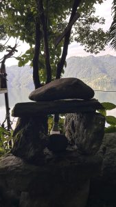 A serene lakeside scene featuring a small stone arrangement topped with a flat rock, surrounded by lush greenery.