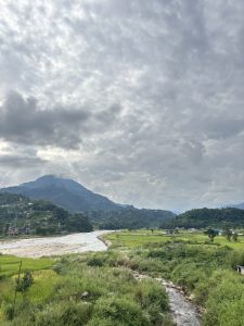 A scenic view of a greenery field and bushes with a small river passing in between, green hills, cloudy sky and small few houses in the hills.
