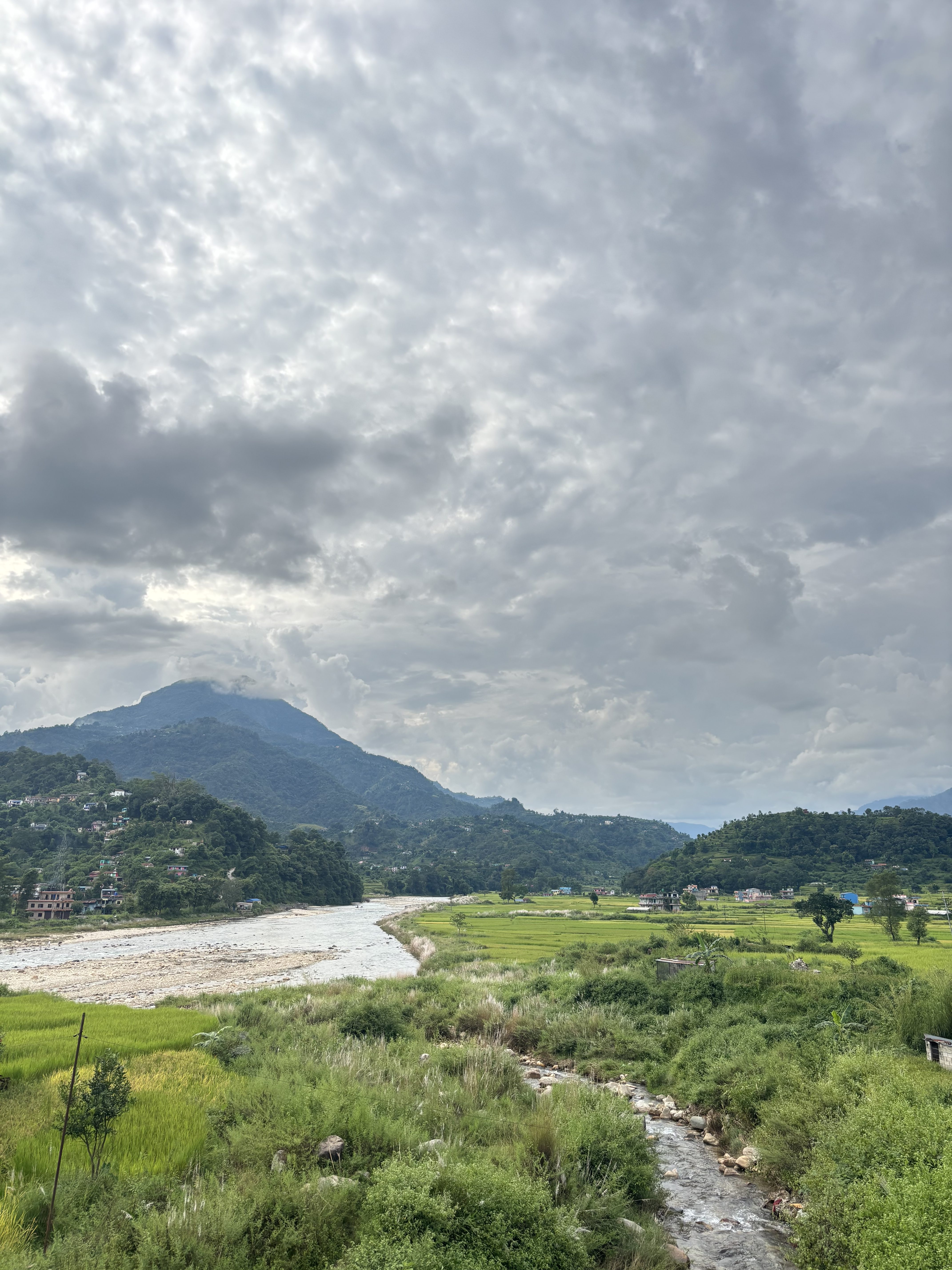 A scenic view of a greenery field and bushes with a small river passing in between, green hills, cloudy sky and small few houses in the hills.