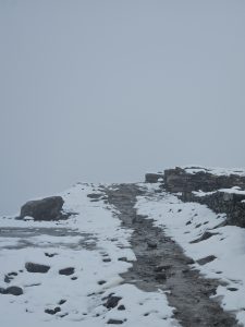  A narrow, uneven path leads through a snowy landscape, flanked by patches of exposed rocks and remnants of stone walls on either side. 
