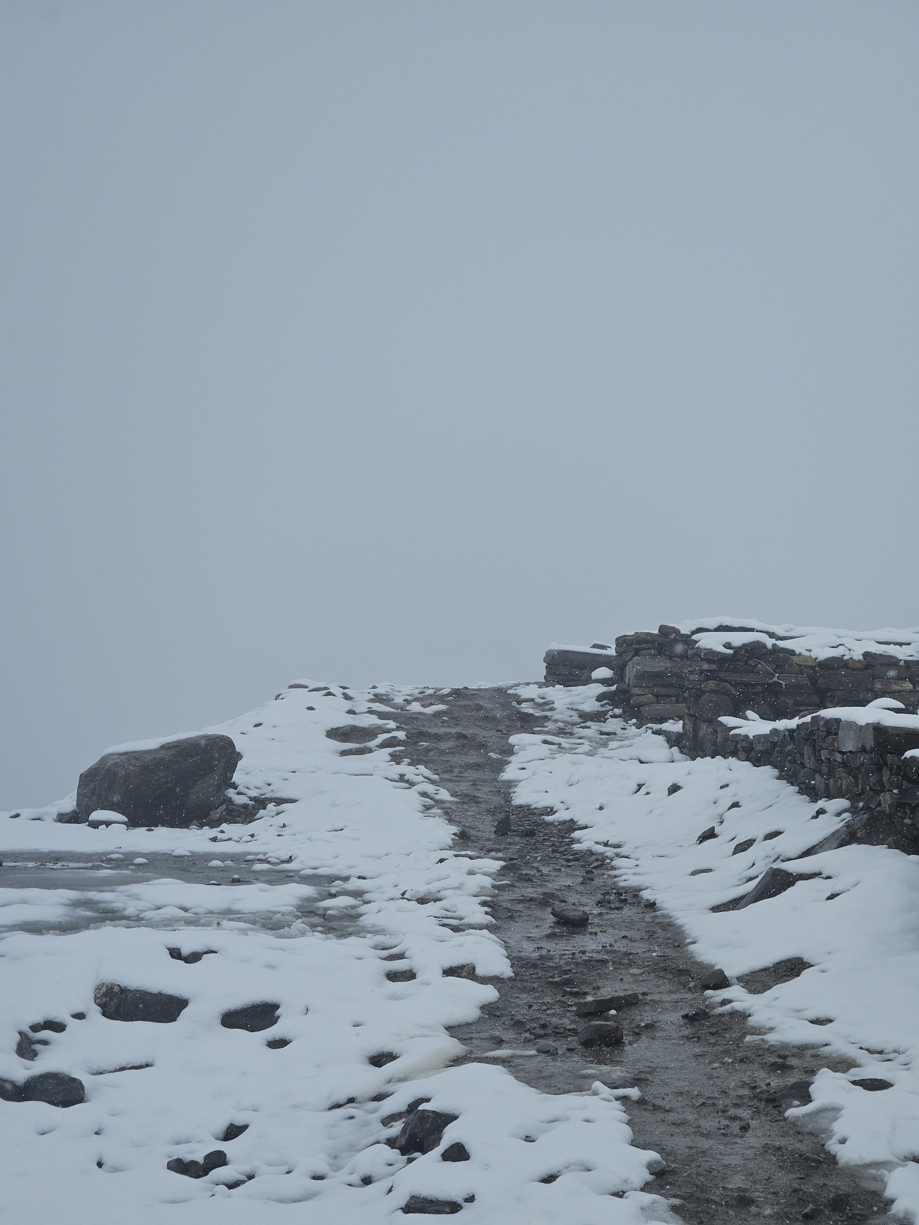 A narrow, uneven path leads through a snowy landscape, flanked by patches of exposed rocks and remnants of stone walls on either side.