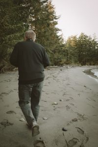 Man walking down a sandy beach with footsteps visible all around and forest trees lining the path.