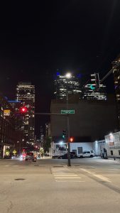 A night scene of an intersection in Chicago. In the center is a tall post with a light at the top, a street sign in the center, and an arm coming off it with a red stop light over the street.