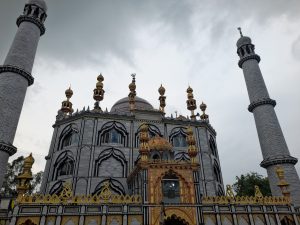 An ornate mosque with two minarets and a grey facade decorated with gold and black patterns under a cloudy sky.