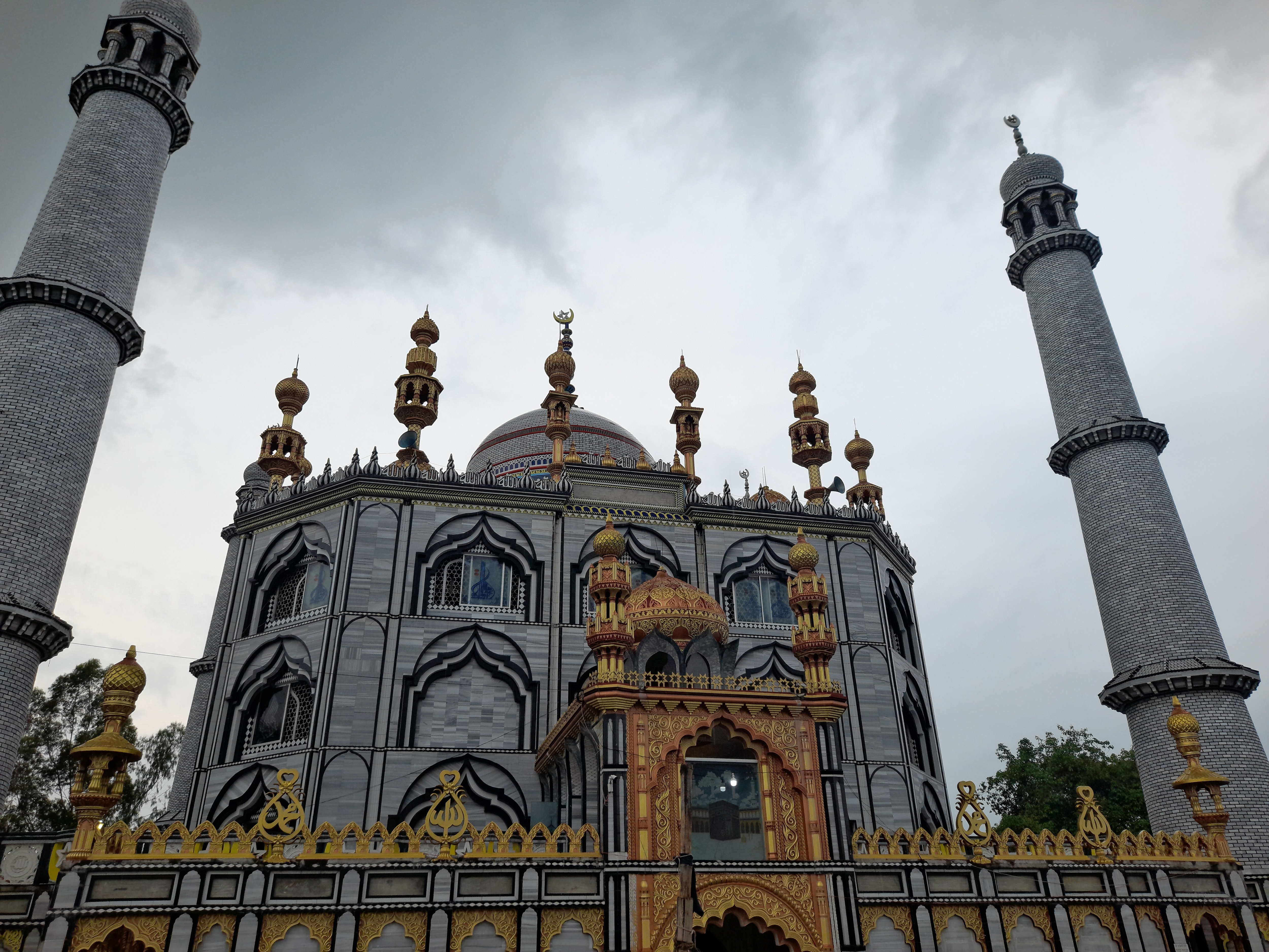 An ornate mosque with two minarets and a grey facade decorated with gold and black patterns under a cloudy sky.