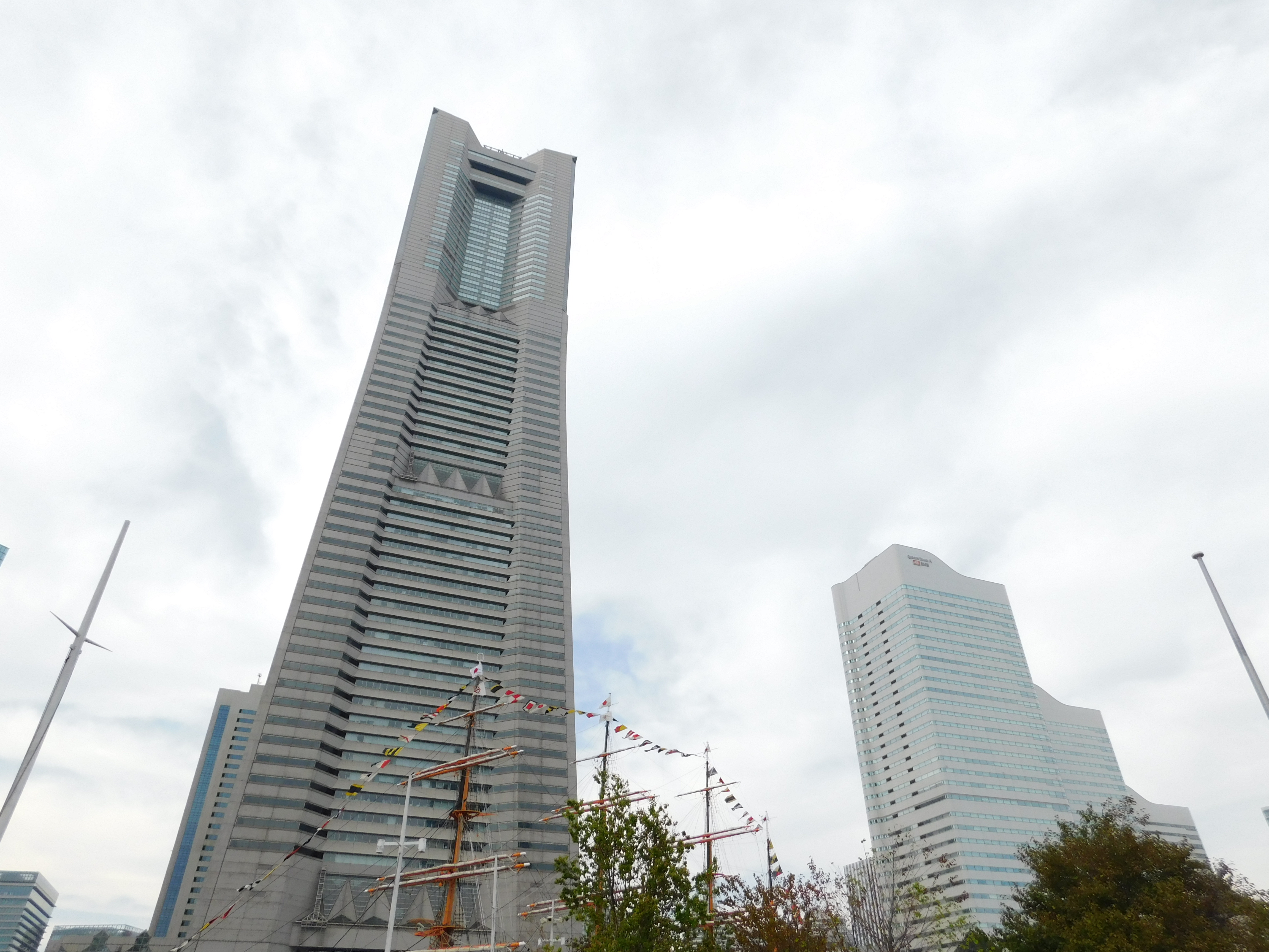 A sleek, modern skyscraper rises against a cloudy sky, its glass and horizontal lines contrasting with a colorful wooden ship in the foreground and mixed-style buildings to the left.