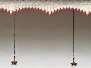 Two traditional hanging brass oil lamps suspended by chains below a decorative roof edge at Sree Shiva Vishnu Temple, Perumanna, Kozhikode. The backdrop is a simple, light-colored wall, highlighting the symmetry and calm of the composition.
