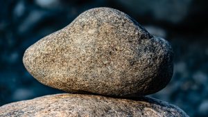 A smooth, rounded stone balances precariously on top of a larger, flat stone. The background is a blurred mix of darker stones and natural surroundings.