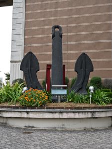 Anchor monument in front of a brown building with visible flowers and greenery.