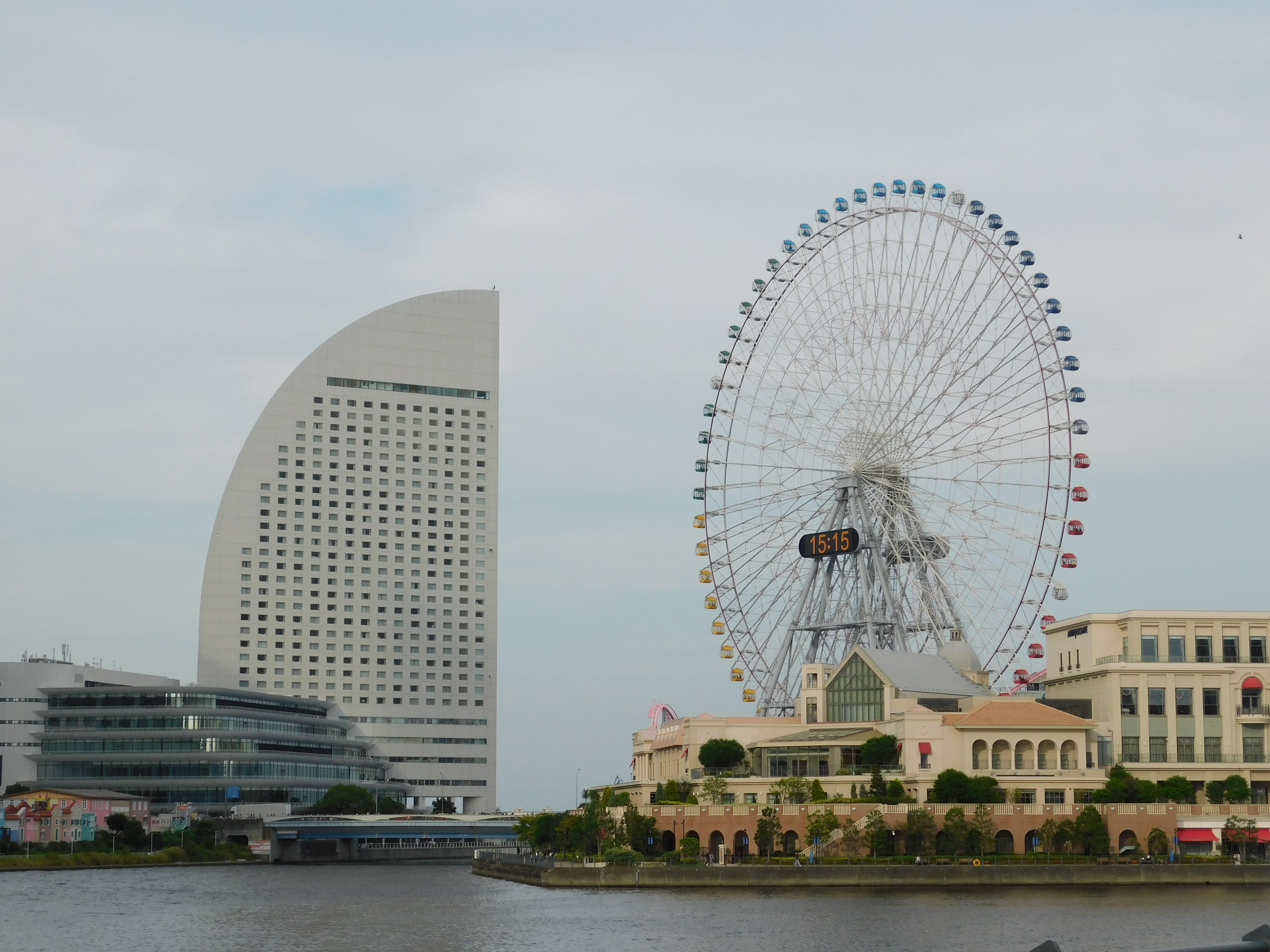 A modern, curved building with a pattern of windows stands on the left side of the image in Yokohama, beside a body of water. To the right, a large Ferris wheel with colorful gondolas displays the time at 15:15. A landscaped area and a building with a white facade occupy the foreground, creating a picturesque urban scene under a cloudy sky.