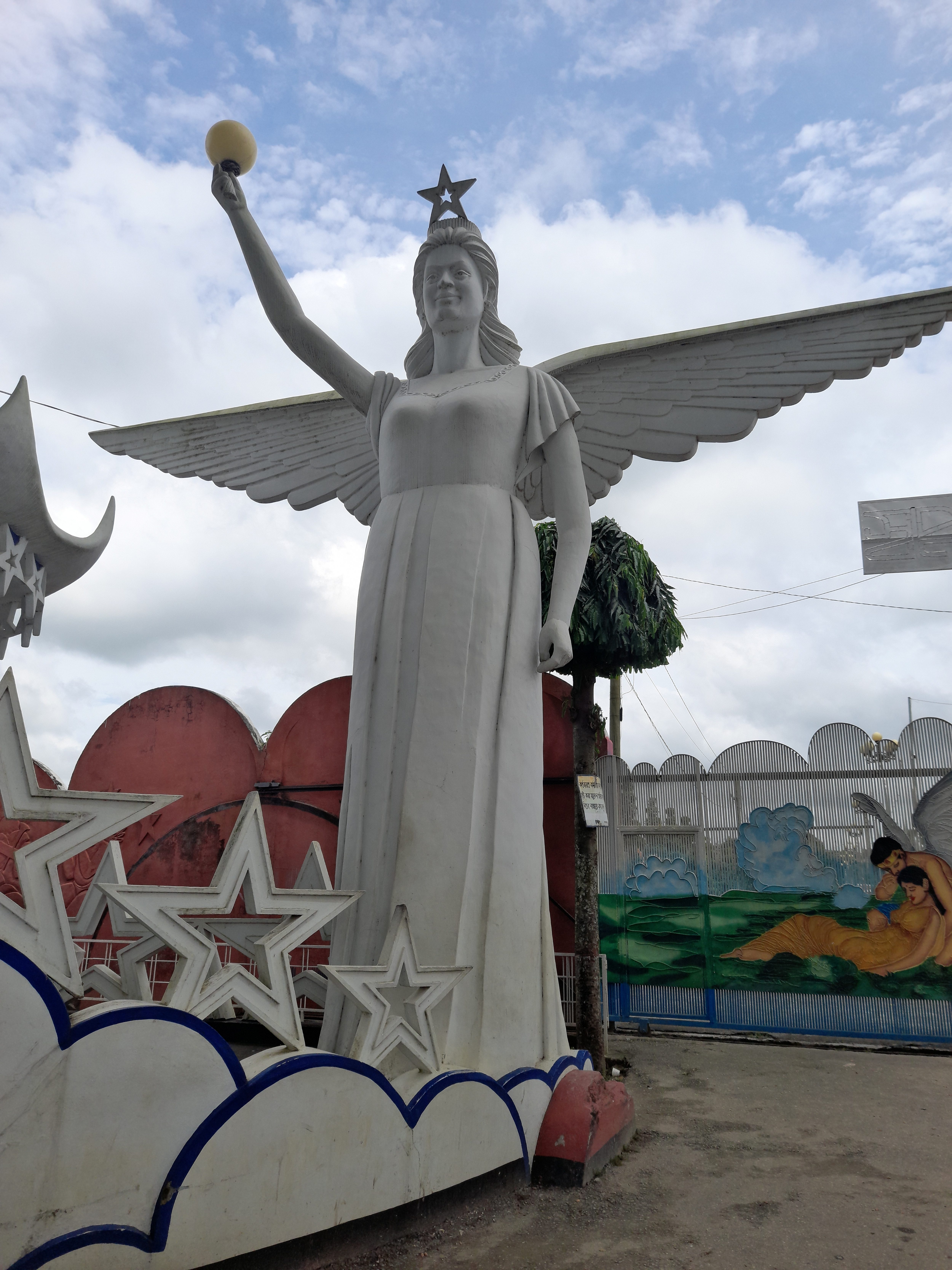 A large, white statue of a woman with wings, dressed in a flowing gown, stands confidently with her right arm raised, holding a spherical object. The figure has a star atop her head and is surrounded by stylized stars and clouds at the base.