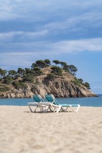 Two empty sun loungers on a sandy beach facing the turquoise sea and a rocky hill with pine trees under a soft blue sky.