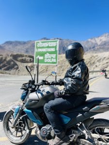 A person sits on racing style bike, seen from the left side. The person is wearing all black, including the helmet.  They&#039;re looking at the camera, but the visor obscures the face.  In the background are mountains in the distance.  The rider sits beside a road side showing directions for Lomanthang and Dolpa in both English and Nepali.