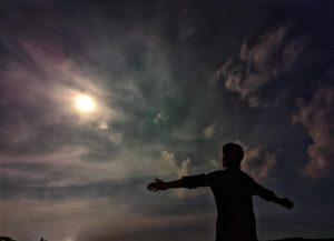 A boy stands with open arms beneath the glowing moon &mdash; as if greeting the sky itself. The night is quiet, the clouds drift slow, and in that soft silver light, he finds a sense of calm only the moon can give.