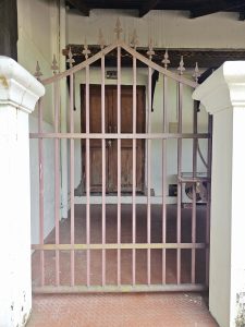 An old iron gate with pointed spikes stands before a wooden door inside a heritage building at Hill Palace, Thrippunithura. The scene shows the charm of traditional Kerala architecture. 