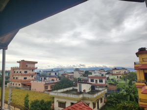 A view from a roof of a house with visible colorful houses, cloudy sky, green paddy field and green hills.