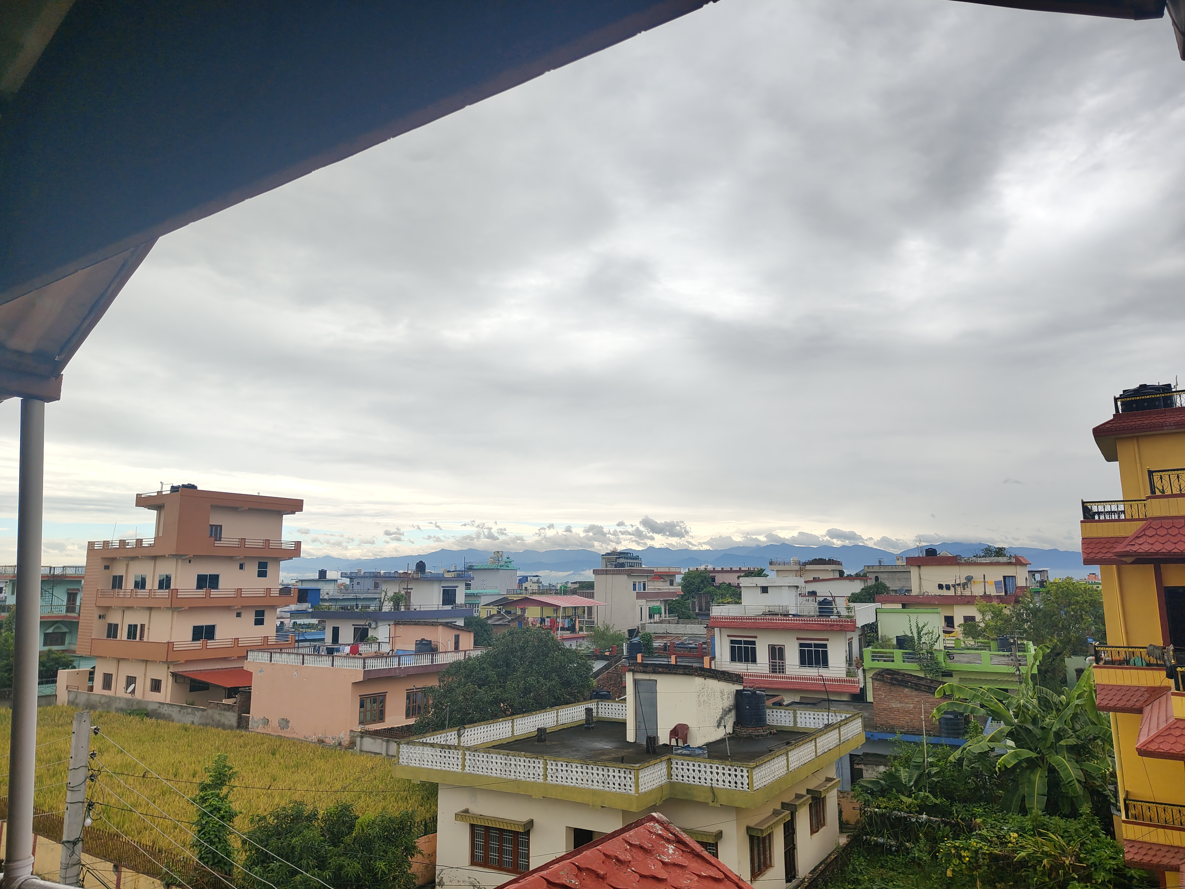 A view from a roof of a house with visible colorful houses, cloudy sky, green paddy field and green hills.