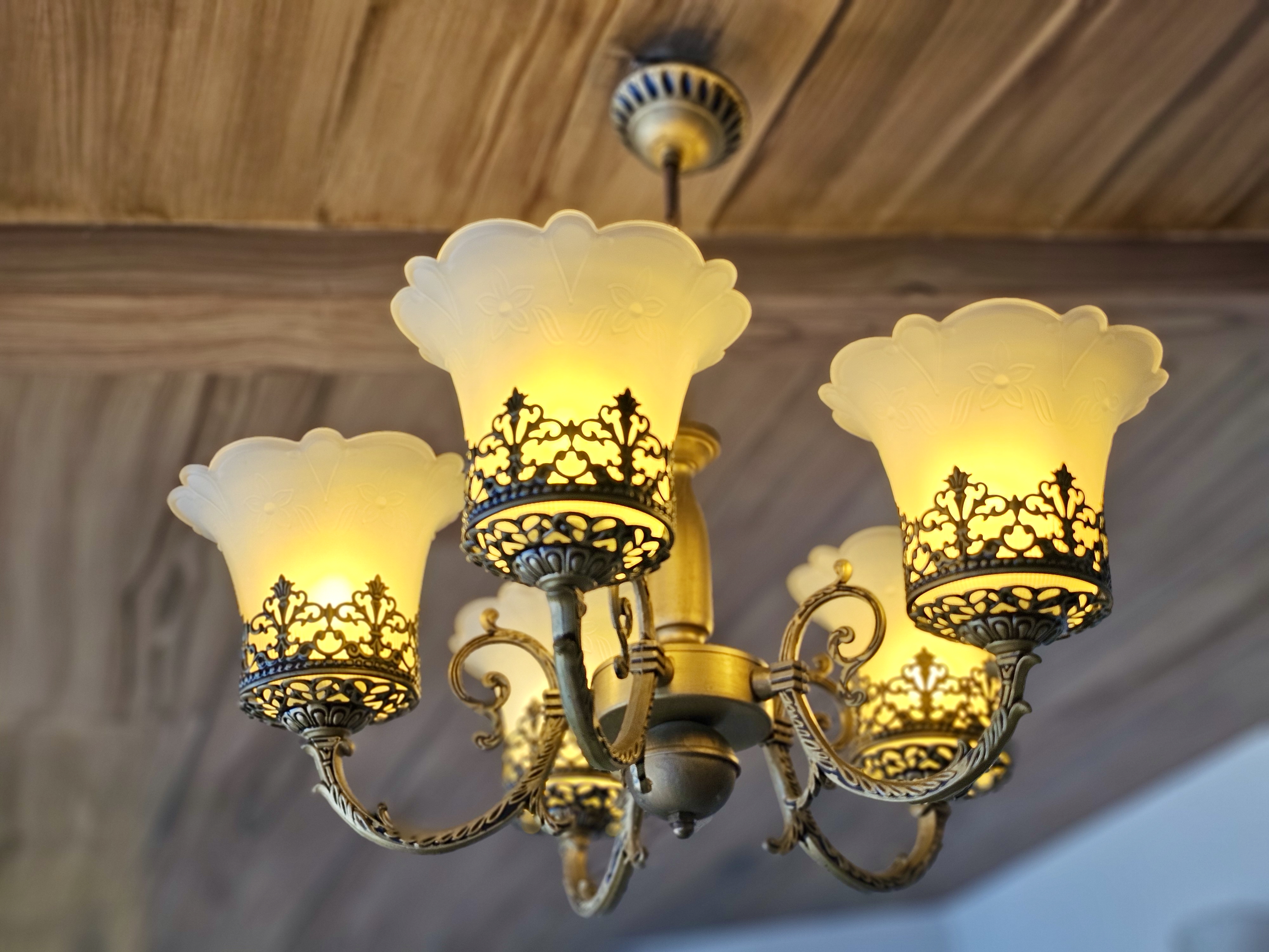 An antique-style chandelier with six warm yellow glass lampshades. The floral designs on the metal and glass add an old-world charm. Shot indoors near Fort Kochi Beach, Kerala.
