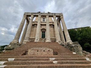 The grand front view of the Temple of Antoninus and Faustina, with tall marble columns and brick steps leading up, stands in the Roman Forum, Rome, Italy. 