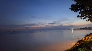 The vast Padma River at twilight, showing the reflective water and the silhouetted riverbank with distant city lights glowing under a blue and purple gradient sky.
