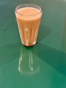 A small glass of steaming Indian chai sits on a green table, reflecting soft light. Photographed at a local tea shop in Palazhi, Kozhikode. 