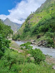 A scenic landscape featuring a lush green hillside with rocky formations, a flowing river along the bottom, and a bright blue sky partially covered by white clouds. 