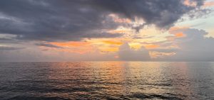 A serene ocean view at sunset, featuring calm waters reflecting shades of orange and pink from the sky. The horizon is lined with soft, fluffy clouds, and the atmosphere is a blend of moody gray and warm colors, creating a tranquil scene.