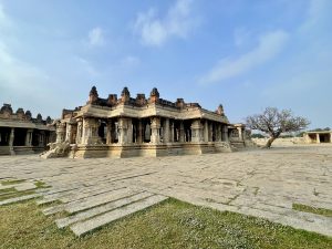 A wide view of the beautifully carved Shree Vijay Vitthala Temple in Hampi, Karnataka, with a lone tree nearby. The image highlights the elegance of Vijayanagara-style architecture under soft daylight. 