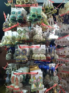 A market stall display of various colorful jhumka earrings hanging on white cardboard tags attached to a metal rack with red strings.