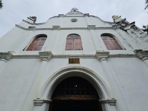 Upward view of the historic white facade of St. Francis Church, Fort Kochi, Kerala, with three wooden windows and an arched doorway. It is one of the oldest European churches in India. 