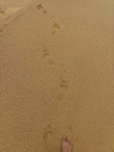 A close-up view of sandy beach terrain with a single bare foot positioned in the foreground. Several footprints are visible trailing off into the distance, leaving impressions in the fine, golden sand.
