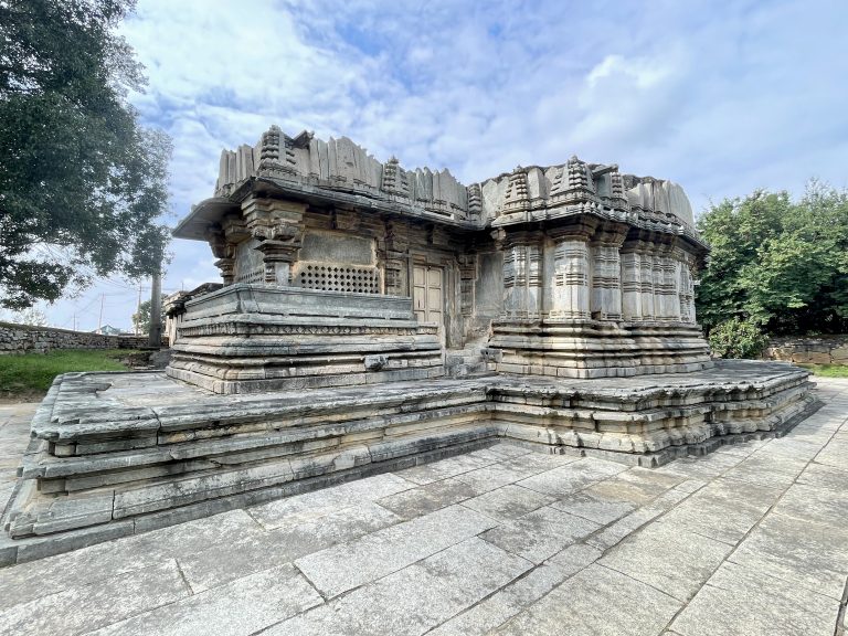 Side view of the temple's back portion at Lakshmi Narasimha Temple, Nuggehalli, Hassan. The stone platform and rooflines stand out against a partly cloudy sky and trees.