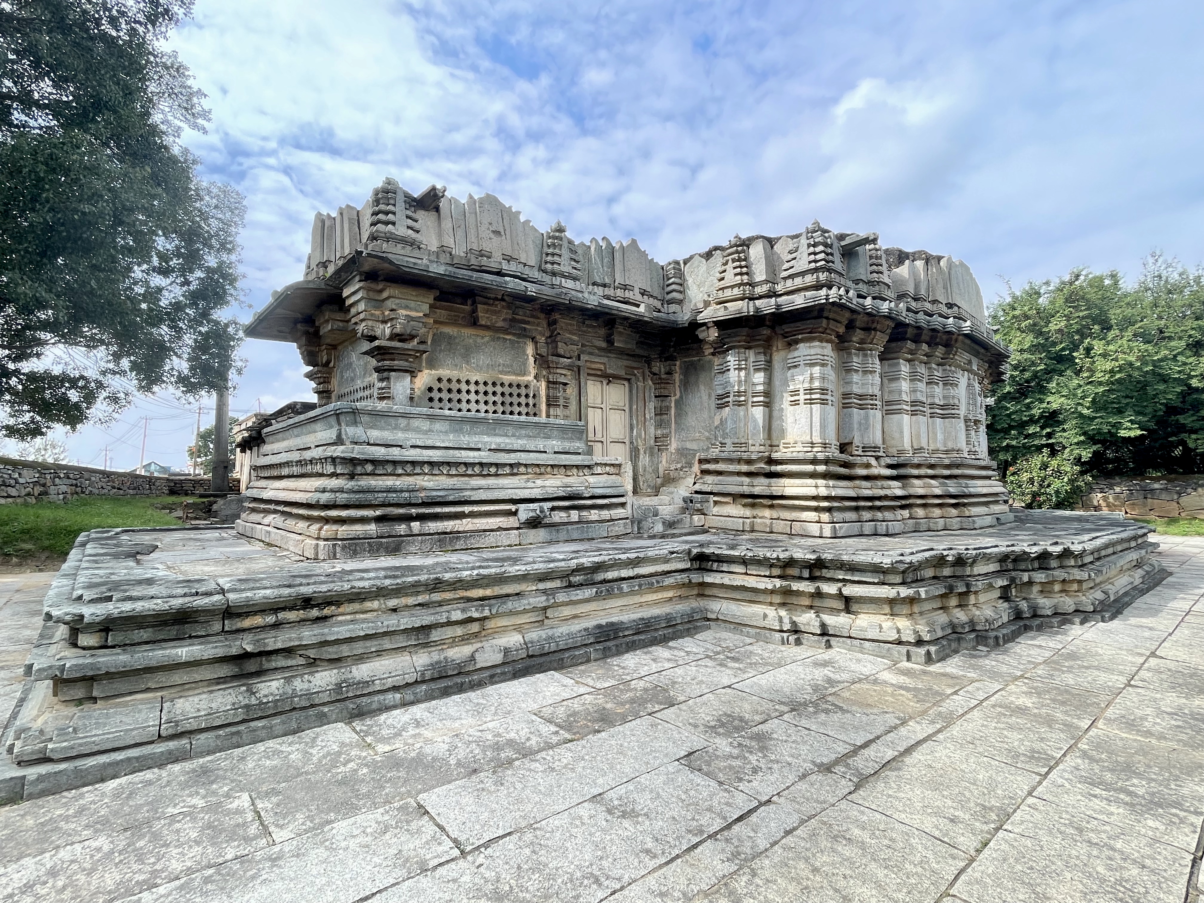 Side view of the temple's back portion at Lakshmi Narasimha Temple, Nuggehalli, Hassan. The stone platform and rooflines stand out against a partly cloudy sky and trees. 