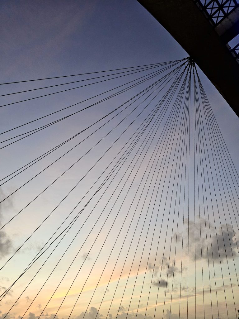 Thin steel bridge cables stretch from a high point, forming symmetrical lines across a soft pastel evening sky. Taken at Marine Drive, Kochi.