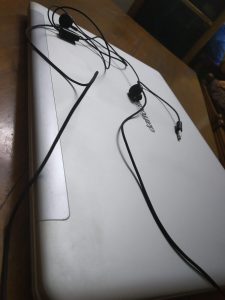 A high-angle close-up of a pair of tangled black wired earphones lying on a closed, silver-gray lid of a laptop. The laptop is resting on a wooden table. 
