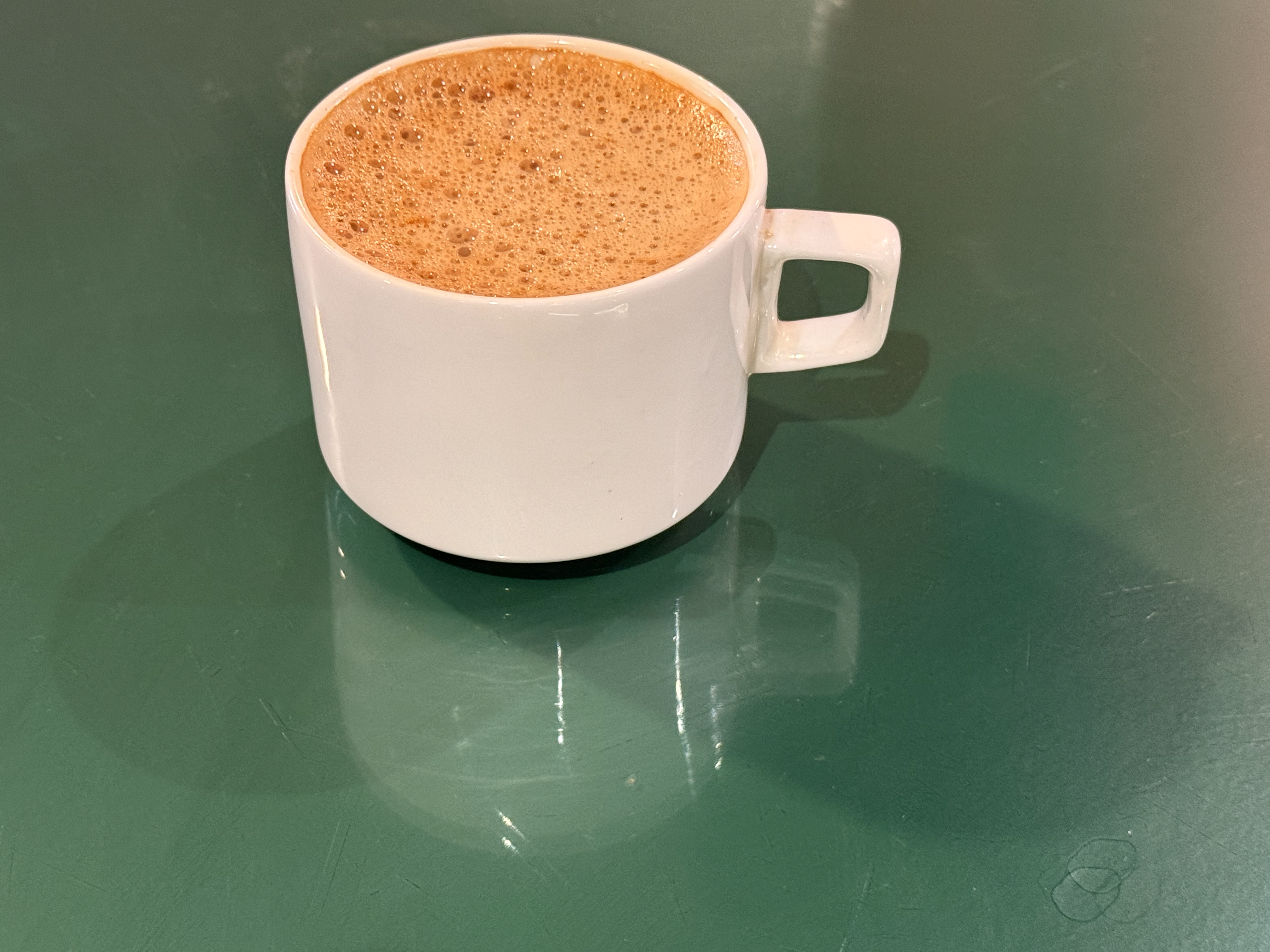 A close-up of a white ceramic coffee cup filled with frothy filter coffee, placed on a shiny green tabletop. Captured indoors at a local cafe in Palazhi, Kozhikode. 