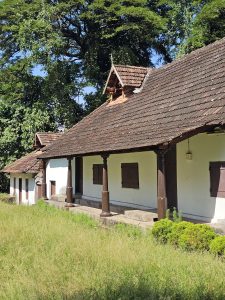 A traditional Kerala-style building with white walls, a brown tiled roof, and wooden columns. Surrounded by greenery and bright sunlight, captured at Hill Palace, Thrippunithura, Kerala.