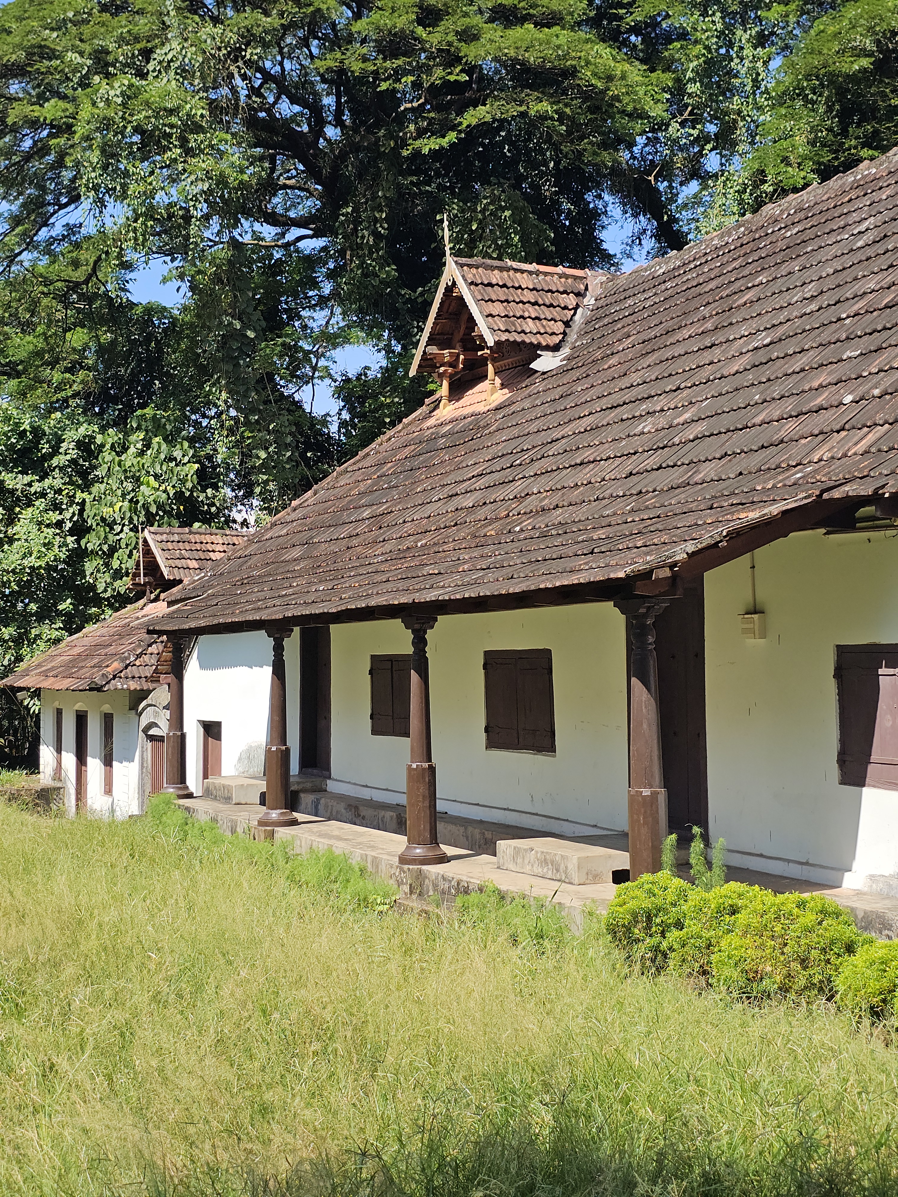 A traditional Kerala-style building with white walls, a brown tiled roof, and wooden columns. Surrounded by greenery and bright sunlight, captured at Hill Palace, Thrippunithura, Kerala.