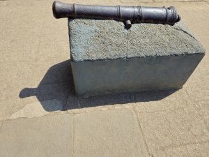 An old iron cannon placed on a rectangular stone pedestal at Shaniwar Wada, Pune, Maharashtra. The bright sunlight creates a sharp shadow of the cannon on the stone-paved ground, highlighting its historic structure.