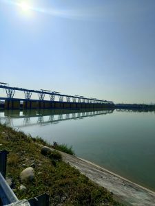 A calm water body reflects a clear blue sky under bright sunlight. On one side, there is a long structure with yellow and blue segments, possibly a dam or a water management facility.