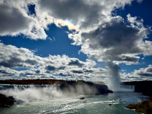 A wide daytime view of Niagara Falls under a partly cloudy sky, with mist rising dramatically from the cascading water below. The sunlight filters through the clouds, casting a silvery glow on the river’s surface, while a tour boat approaches the falls surrounded by the natural beauty of the scene.
