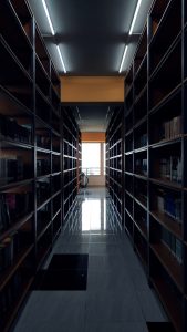 A book library with tons of collections placed on a bookshelf attached to the wall. Visible marble flooring and tubelights on the ceiling.