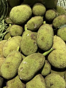 A stack of fresh, tender jackfruits with rough green skin and stem intact, displayed in a market in Palazhi, Kozhikode. Bright sunlight highlights the fruit textures.