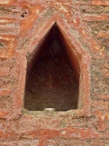 A close-up of a weathered, reddish-orange brick wall featuring a dark, triangular niche. A small, white object rests at the bottom of the alcove. Captured at Parvati Hill, Pune.