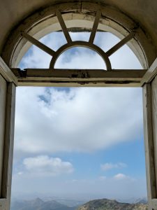 A view through a semi-circular arched window showing distant Aravalli hills and blue sky. Captured inside Monsoon Palace, Sajjangarh, Udaipur.