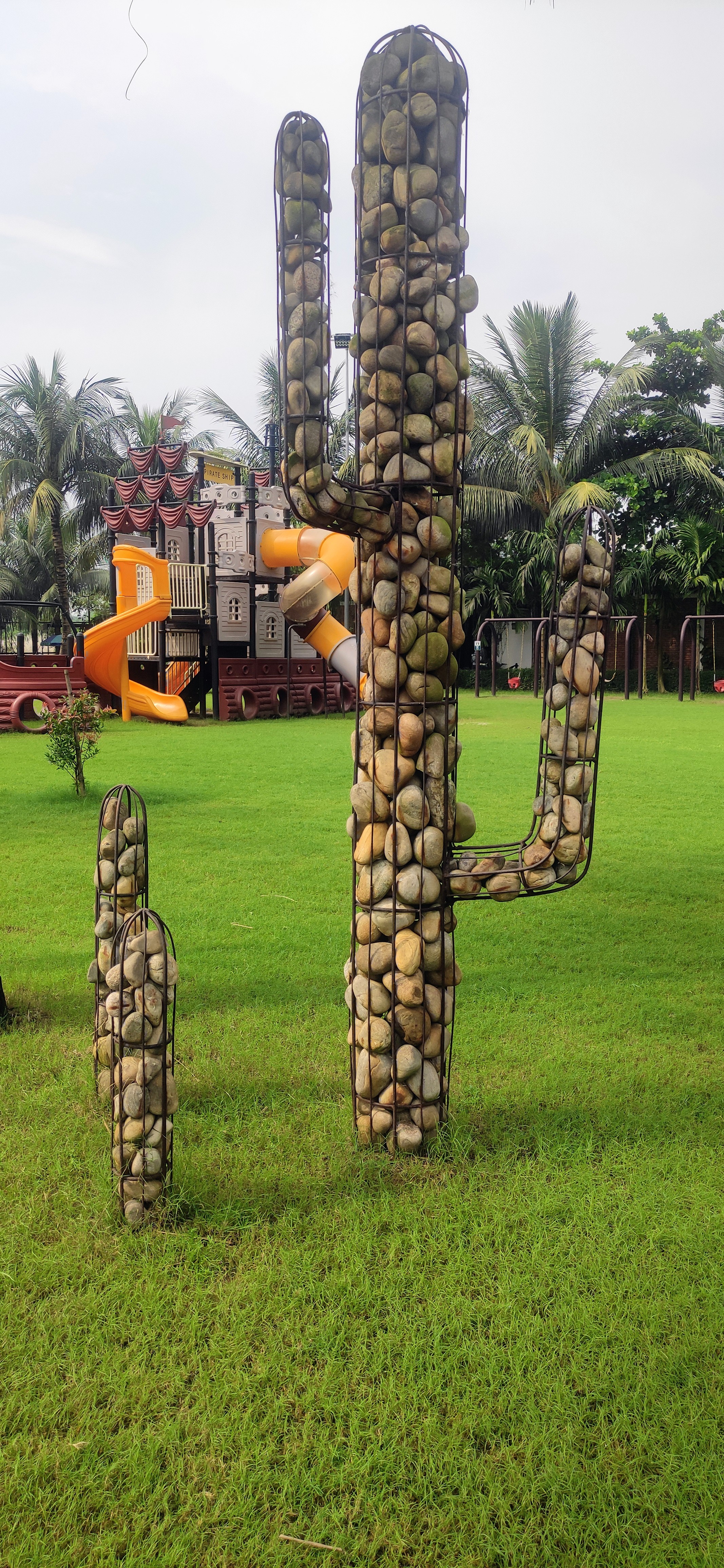 A wire-frame cactus sculpture filled with smooth stones stands on grass, with a colorful playground and palm trees visible in the background under a partly cloudy sky.