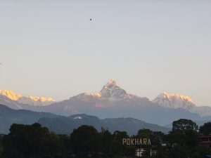 Snow-capped mountains bathed in golden sunlight rise above lush greenery, with a "POKHARA" sign and birds flying in a clear sky.