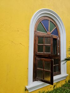 A beautiful wooden arched window with textured stained glass set against a vibrant yellow wall near Fort Kochi Beach. The half-open window adds character to the colonial-style architecture.