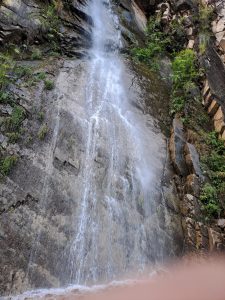A cascading waterfall flows down a rocky cliff face, surrounded by patches of green vegetation.