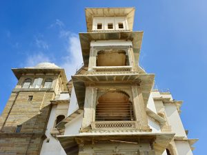 An angled view of the detailed outer facade of Monsoon Palace, Sajjangarh, Udaipur. Features aged balconies, domes, and stone textures against a bright blue sky.